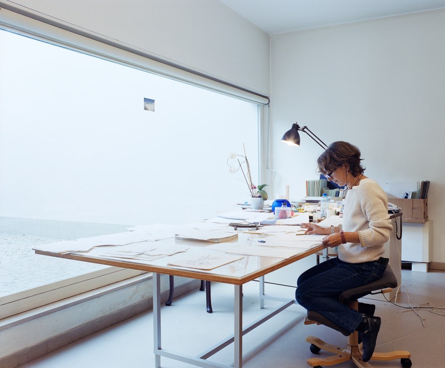 Archivist Chiara Porcu working at a drafting table in a bright, minimalist office with large windows overlooking water.