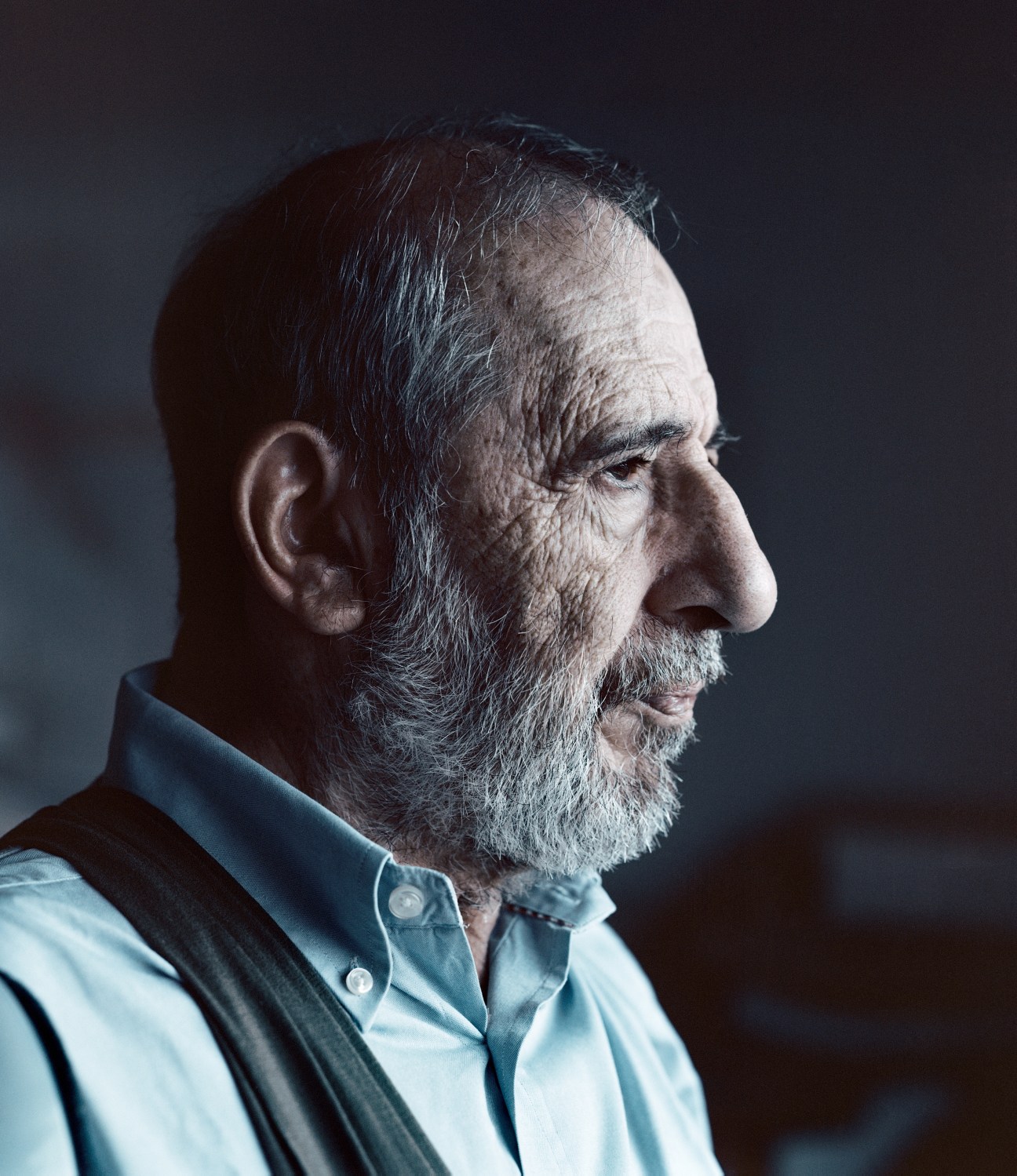 Portrait of elderly man with grey beard wearing light blue shirt against dark background.