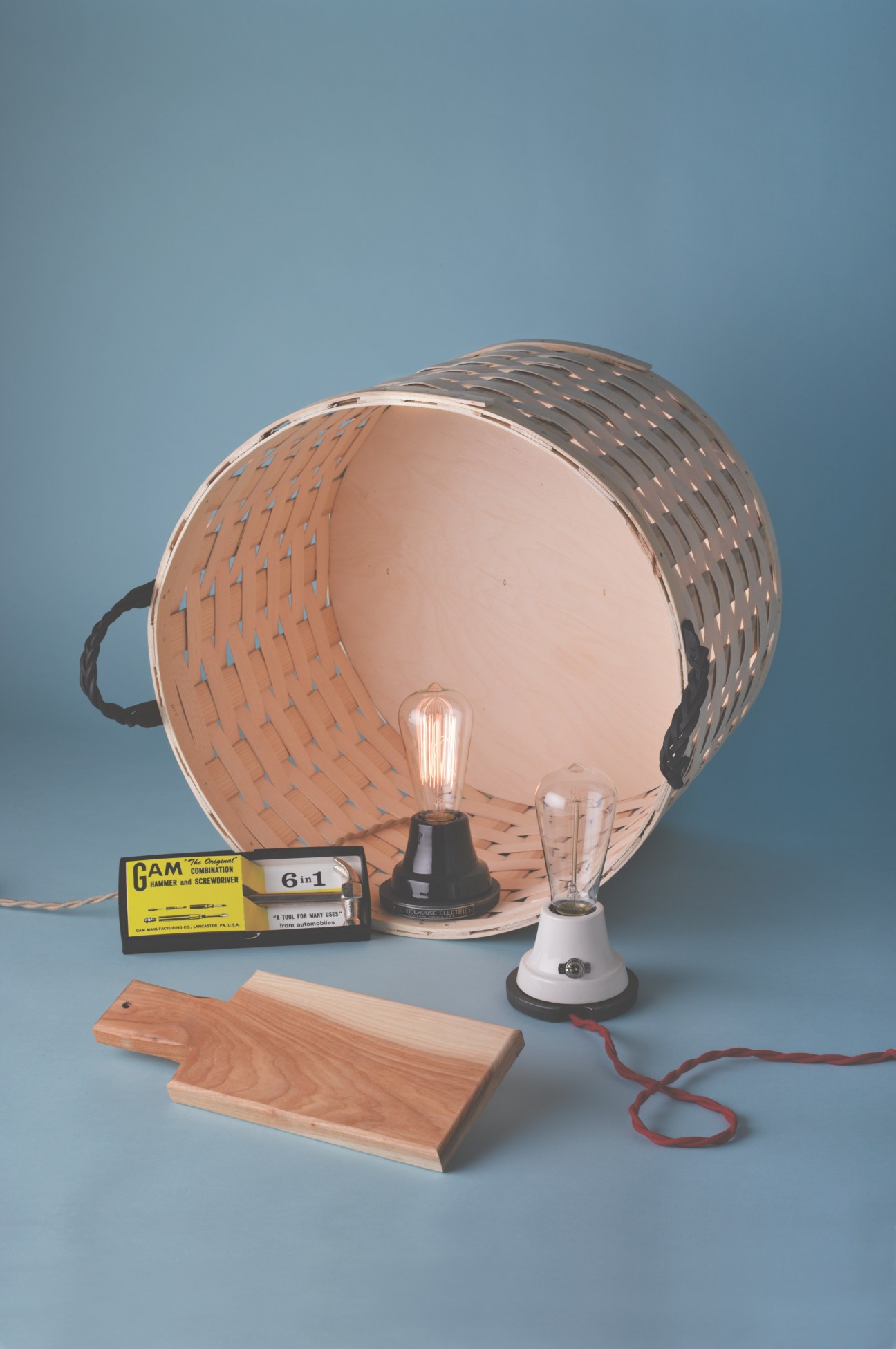 Schoolhouse Electric products including a woven basket lamp, vintage tool, wooden cutting board, and Edison bulb fixture on blue background.