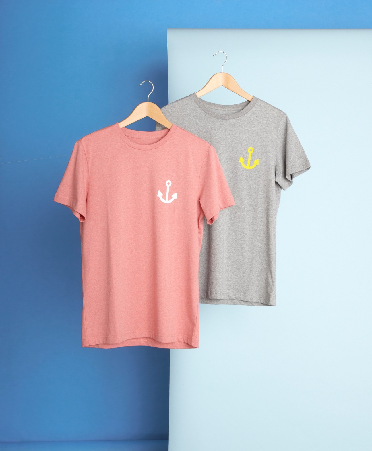 Two cotton T-shirts with anchor prints hanging on wooden hangers against a split blue and white background.