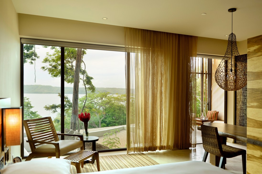 Modern hotel room with floor-to-ceiling windows overlooking a lake and forest, featuring wooden furniture and a woven pendant light.