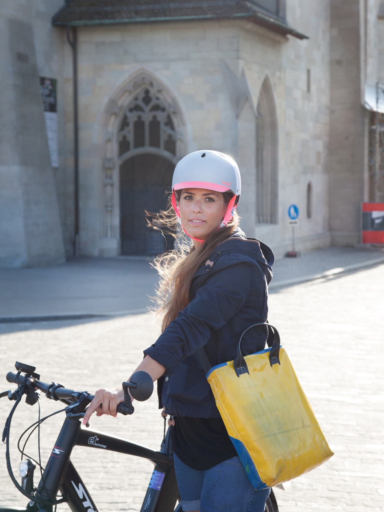 A young woman wearing a white and pink helmet stands with an e-bike in a Zurich square, holding a yellow messenger bag.
