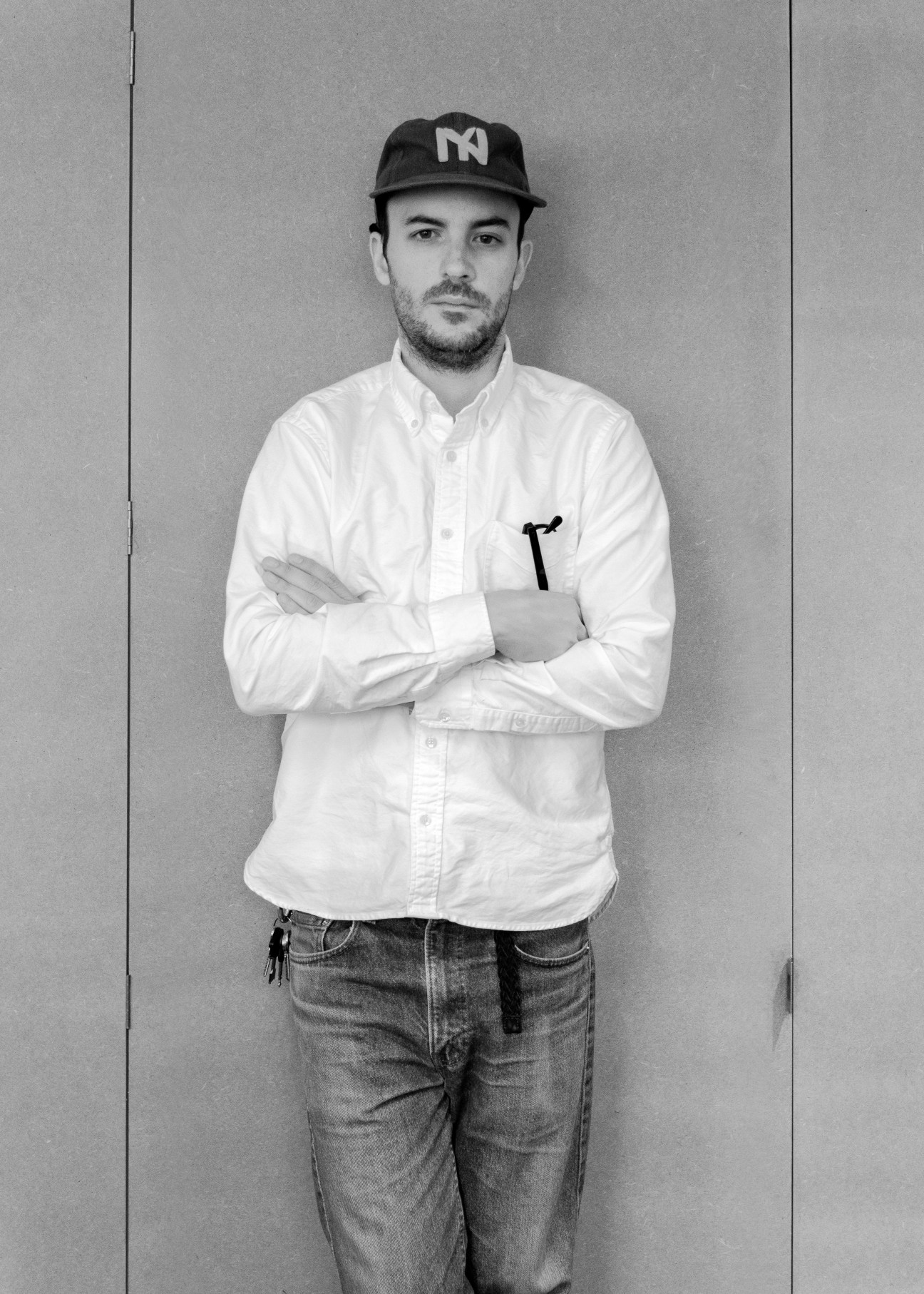 Man wearing white button-up shirt and cap with M logo holds tablet while posing against gray wall.