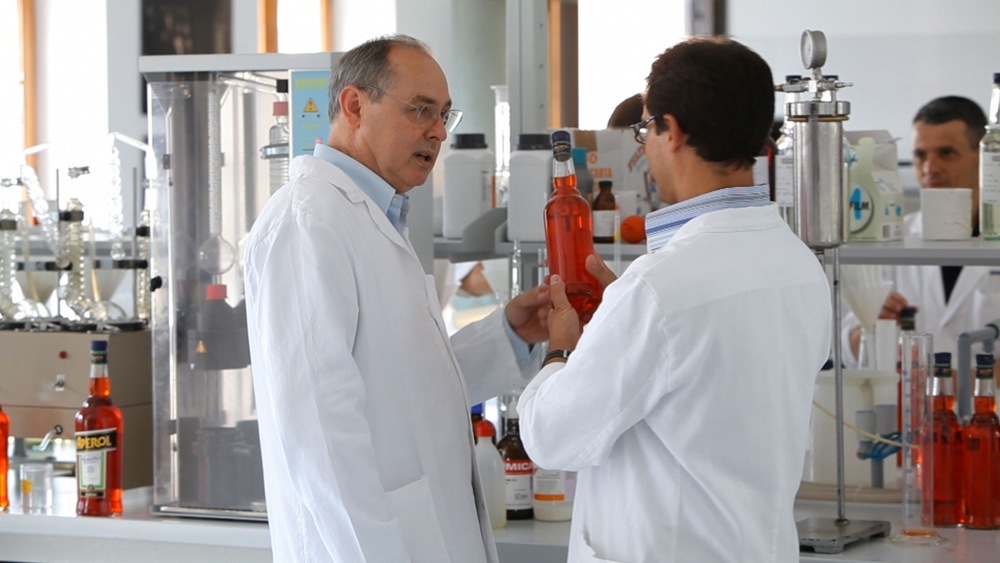 Two men in white lab coats examine an Aperol bottle in a quality control laboratory.