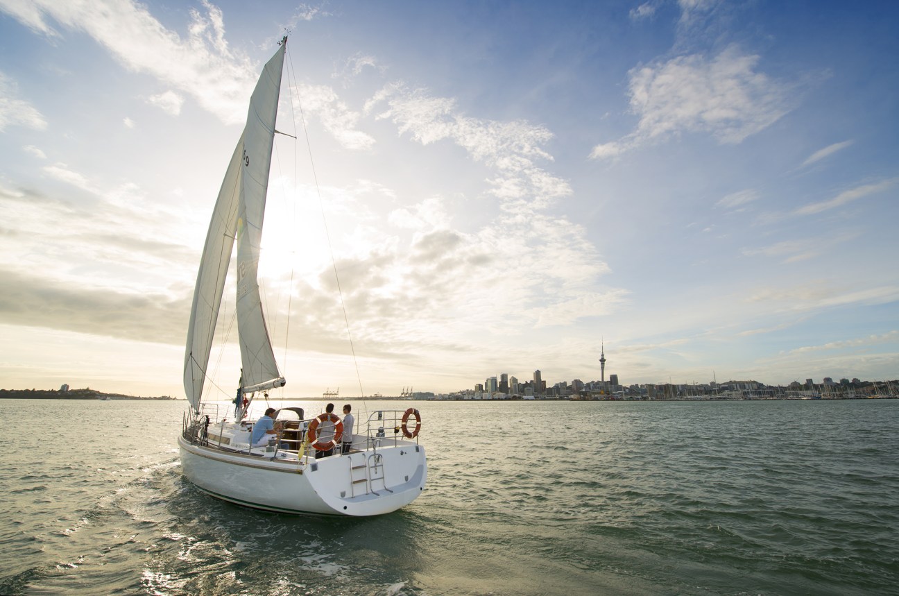 Sailboat with passengers on Auckland harbour with city skyline and Sky Tower in background.