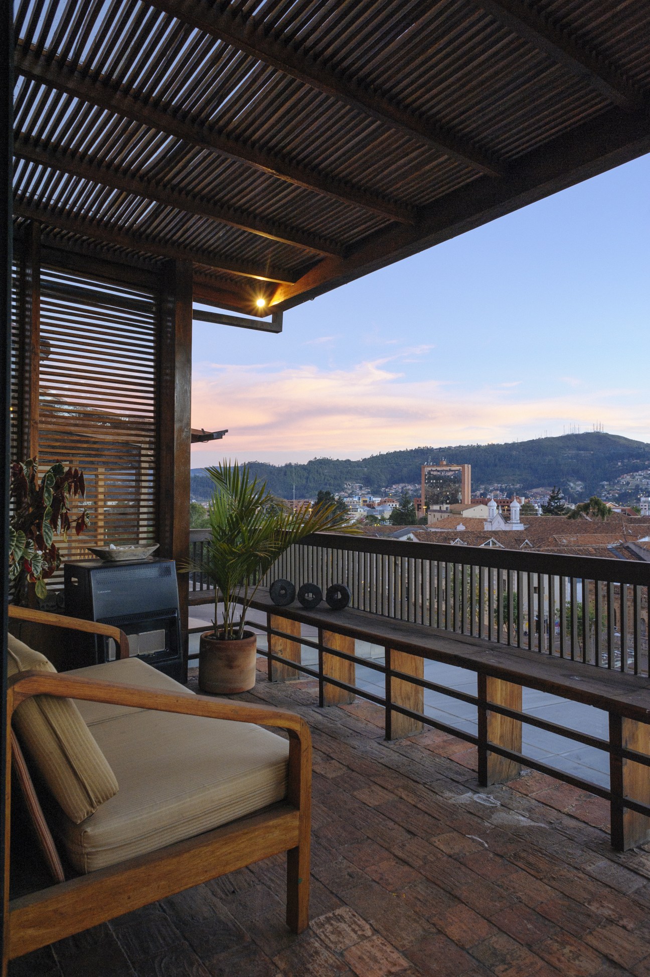 Modern terrace with wooden furniture overlooking Cuenca's colonial cityscape and surrounding mountains at sunset.