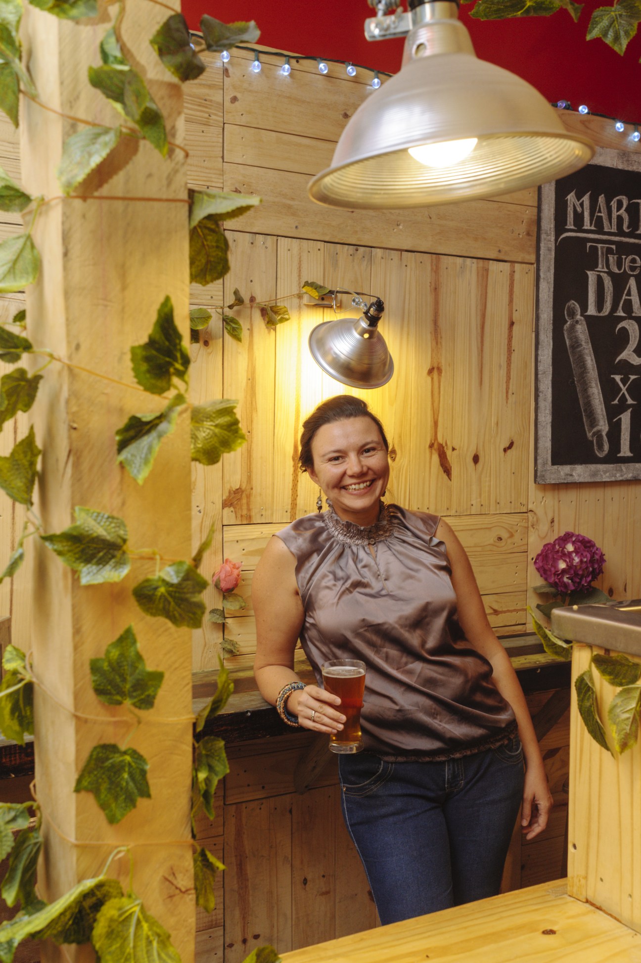 Deborah Debas, owner of Nórdica Brewery, holds a beer glass in her rustic wooden brewery interior decorated with hop plants.