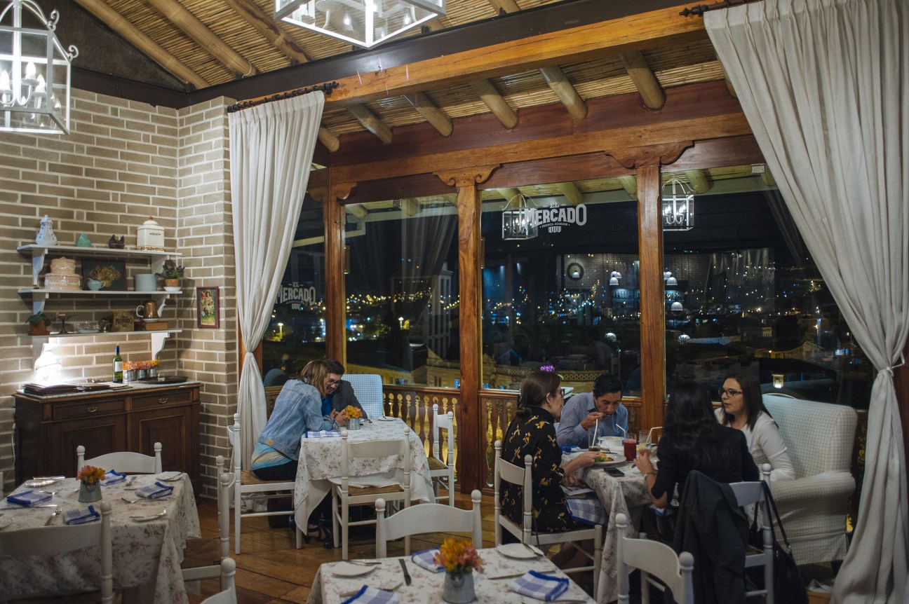 El Mercado restaurant interior with diners seated at tables beside large windows overlooking the city at night.