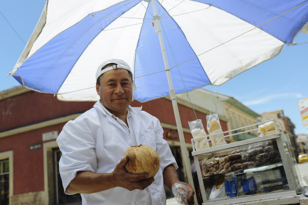 Coconut seller holding fresh coconut under blue and white umbrella at street market stall in Cuenca.
