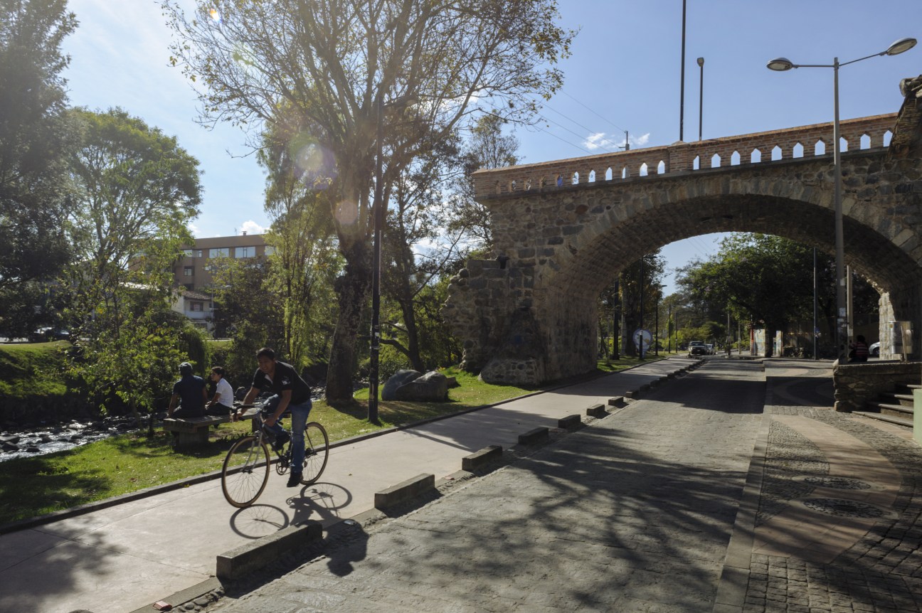 Stone bridge spanning a river in Cuenca with cyclists and pedestrians on a paved path below.