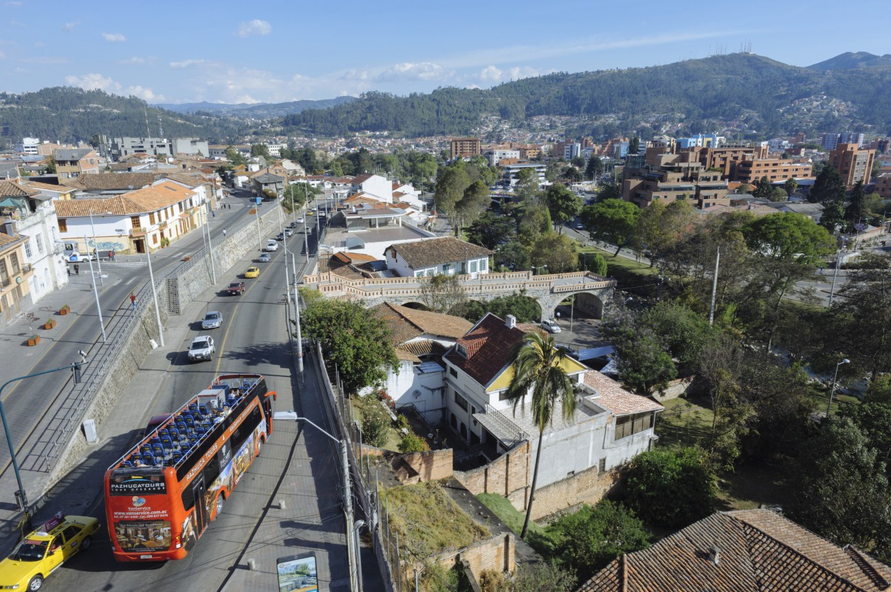 Aerial view of Cuenca's colonial architecture and surrounding Andean mountains with a red tour bus on the street below.