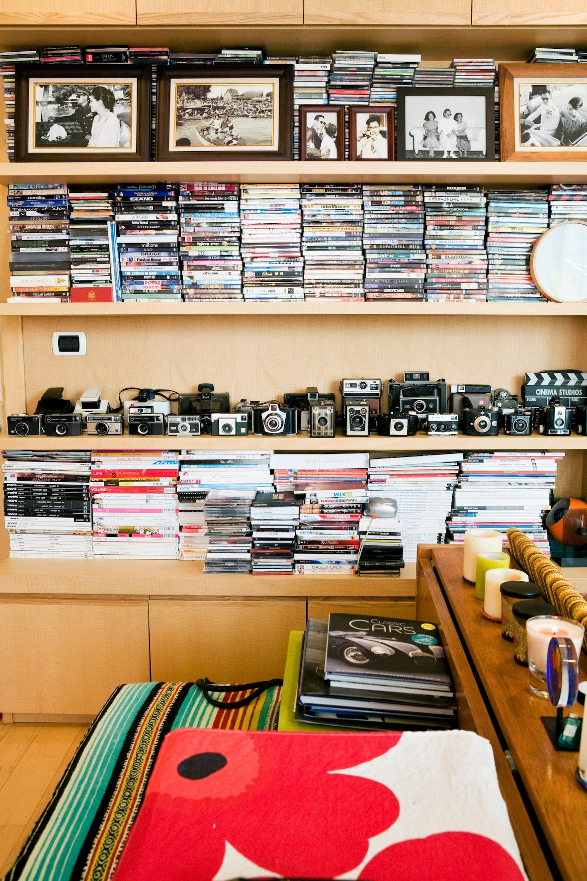 Wooden shelving unit displaying framed vintage photographs, CDs, cameras, magazines, and decorative items including a red and white patterned rug.