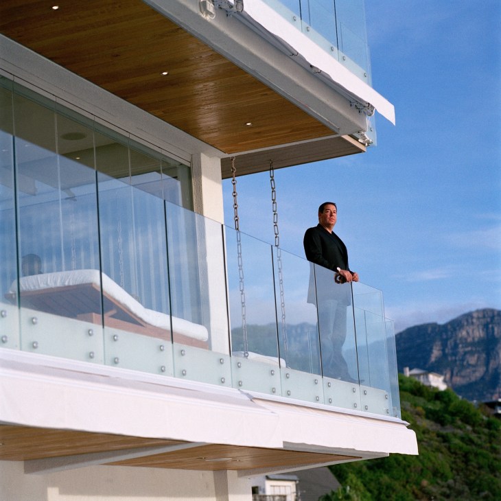 Man in black shirt standing on modern balcony overlooking mountainous landscape.