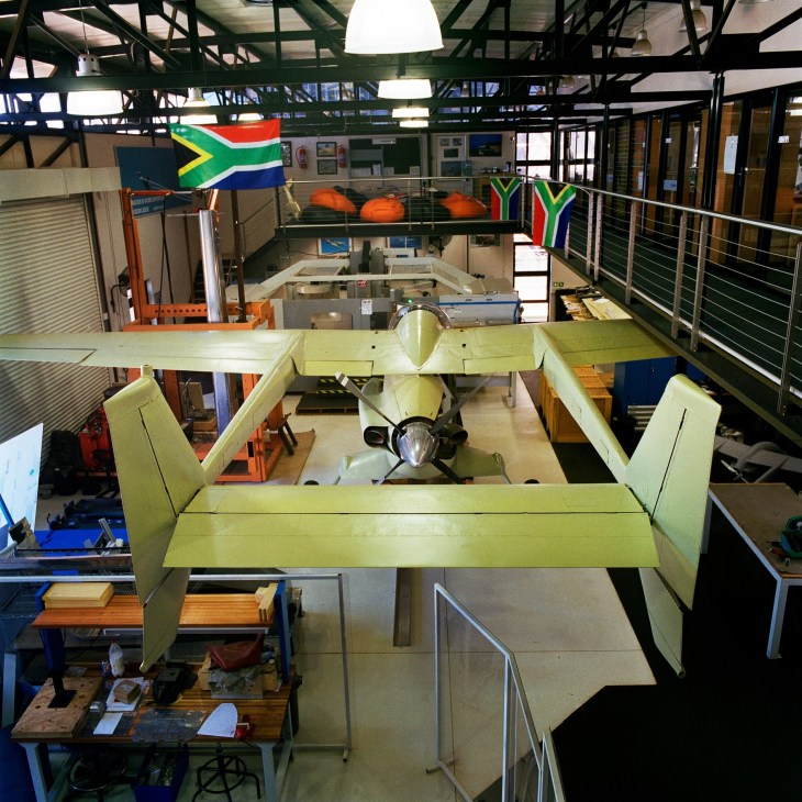 AHRLAC reconnaissance aircraft under construction in a workshop with South African flag displayed overhead.