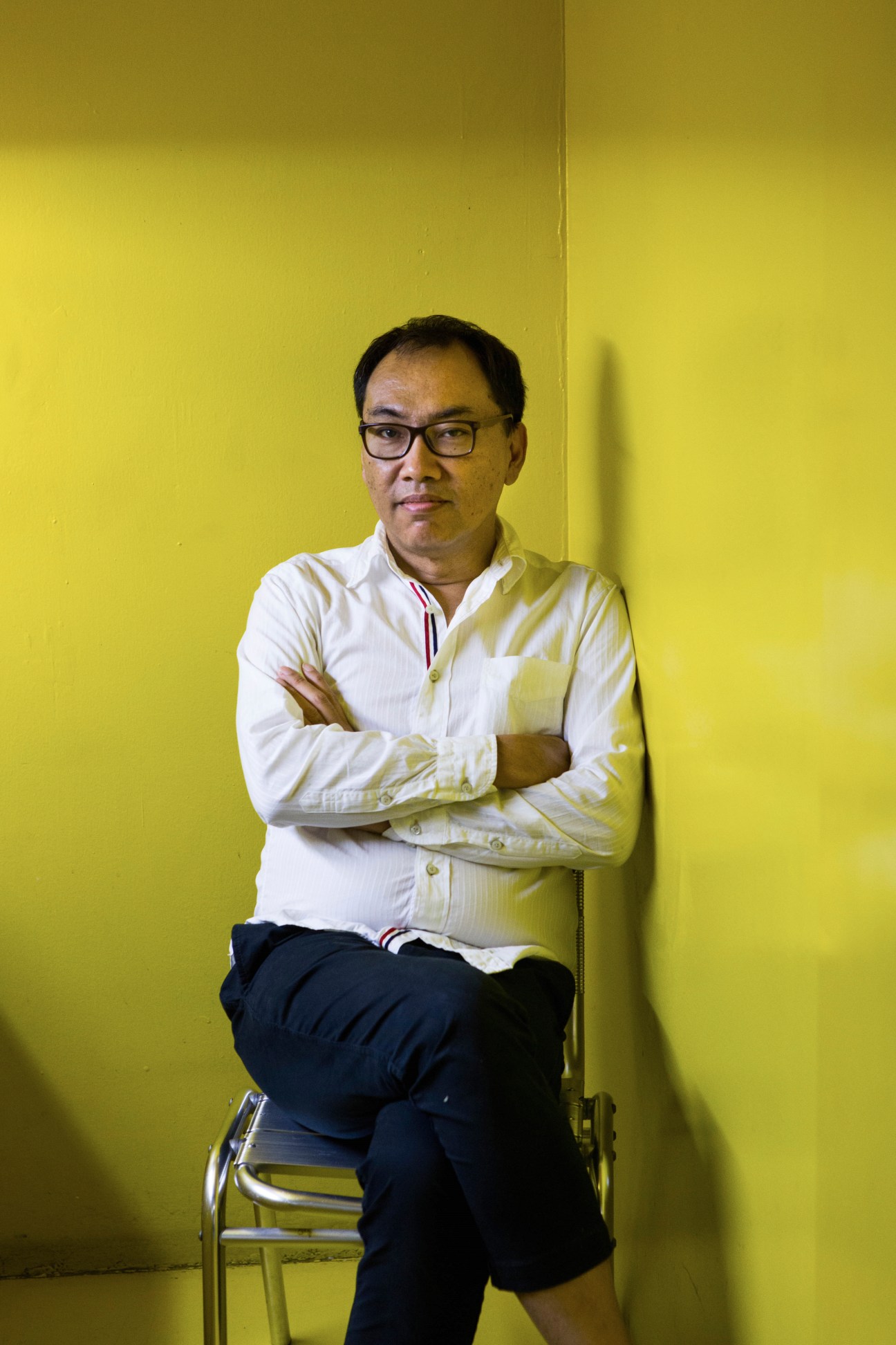 Prartarn Teeratada, editor in chief of Art4d magazine, sitting against a yellow wall wearing glasses
