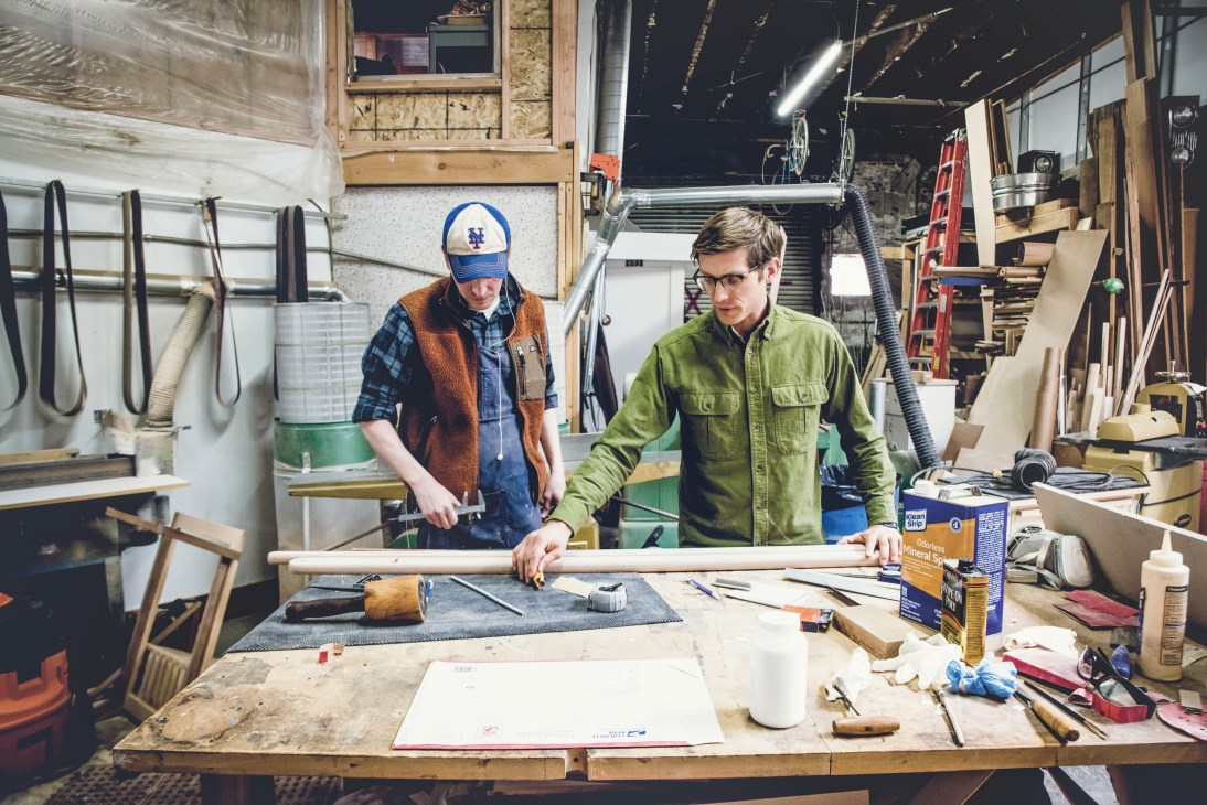 Two craftsmen work at a wooden workbench in Asa Pingree's workshop surrounded by tools and materials.
