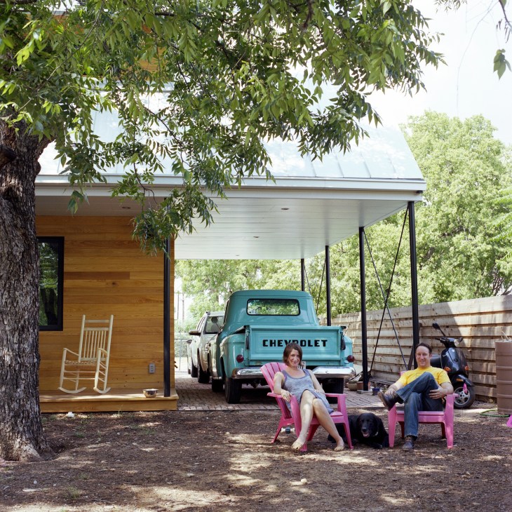 A man and woman sit in pink Adirondack chairs in a backyard with a vintage turquoise Chevrolet truck, wooden cabin, and native trees.