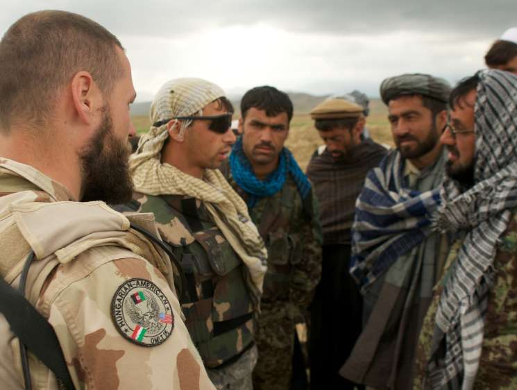 A bearded Western soldier in desert camouflage uniform speaks with Afghan men in traditional clothing in an open field.