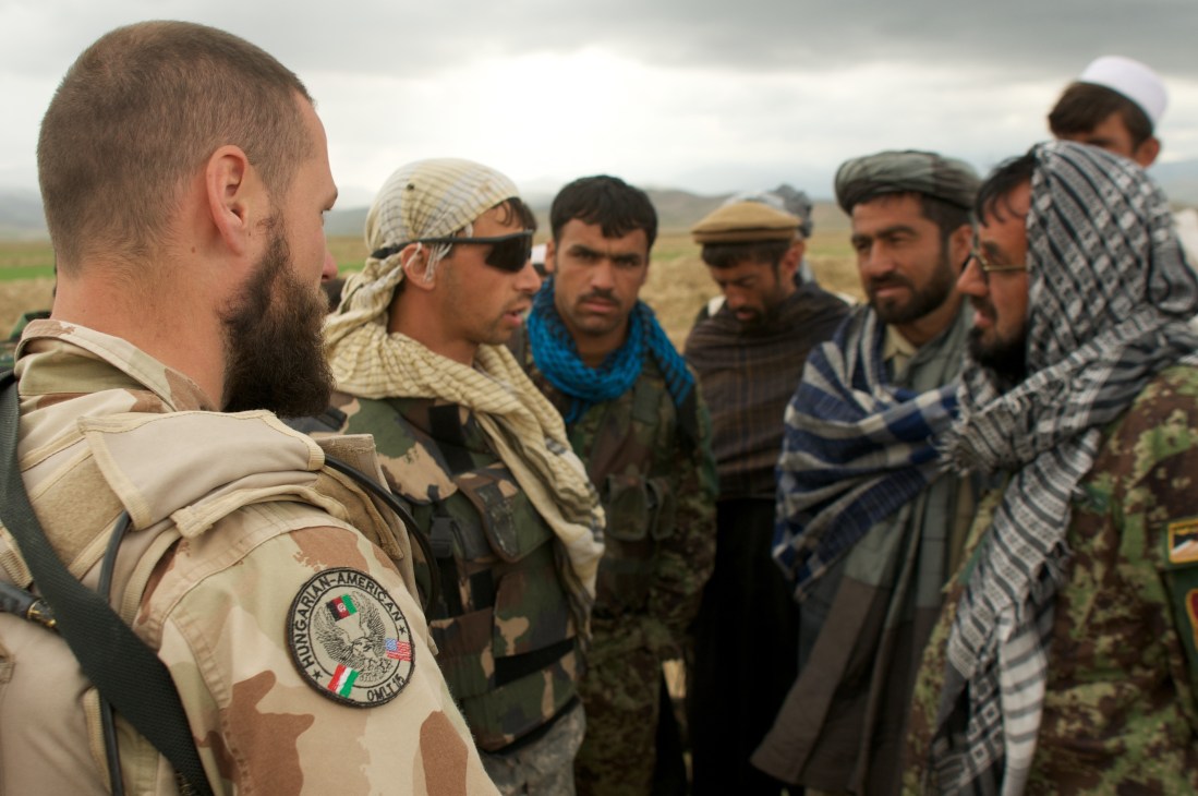 A bearded Western soldier in desert camouflage uniform speaks with Afghan men in traditional clothing in an open field.