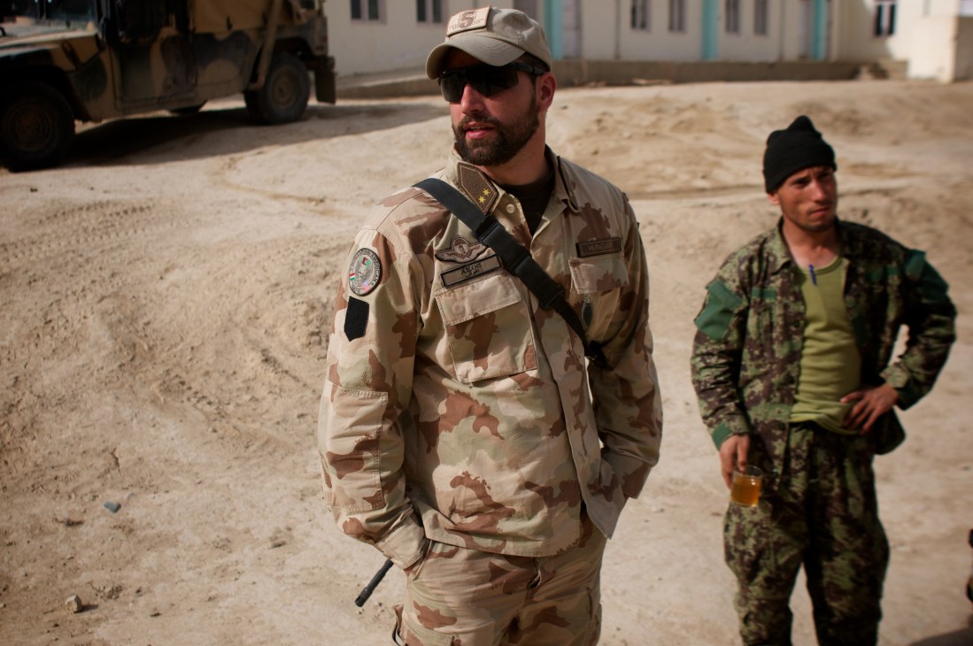 US soldier in desert camouflage uniform stands with Afghan military personnel in a dusty compound in Afghanistan.