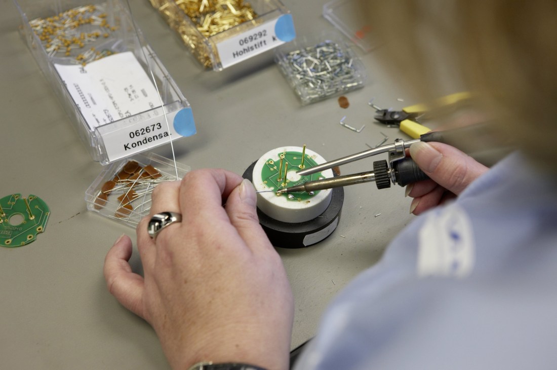 Technician soldering components onto a microphone condenser capsule circuit board during Neumann microphone assembly.