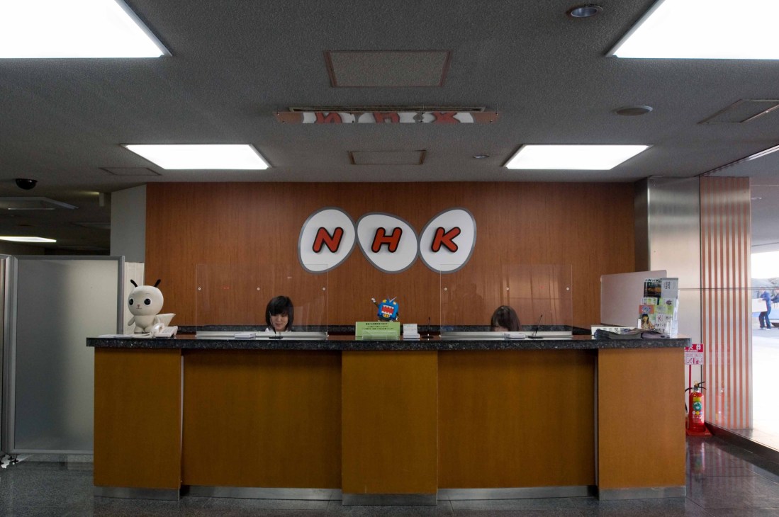 NHK reception desk with staff members and the broadcaster's logo displayed on the wall