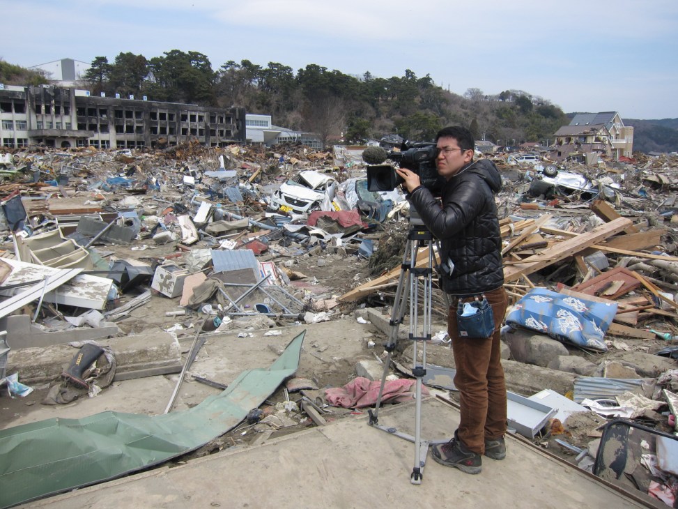NHK cameraman filming tsunami destruction with debris and damaged buildings in background.