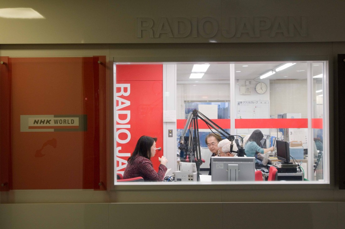 NHK World radio studio with staff working at microphones and broadcasting equipment.