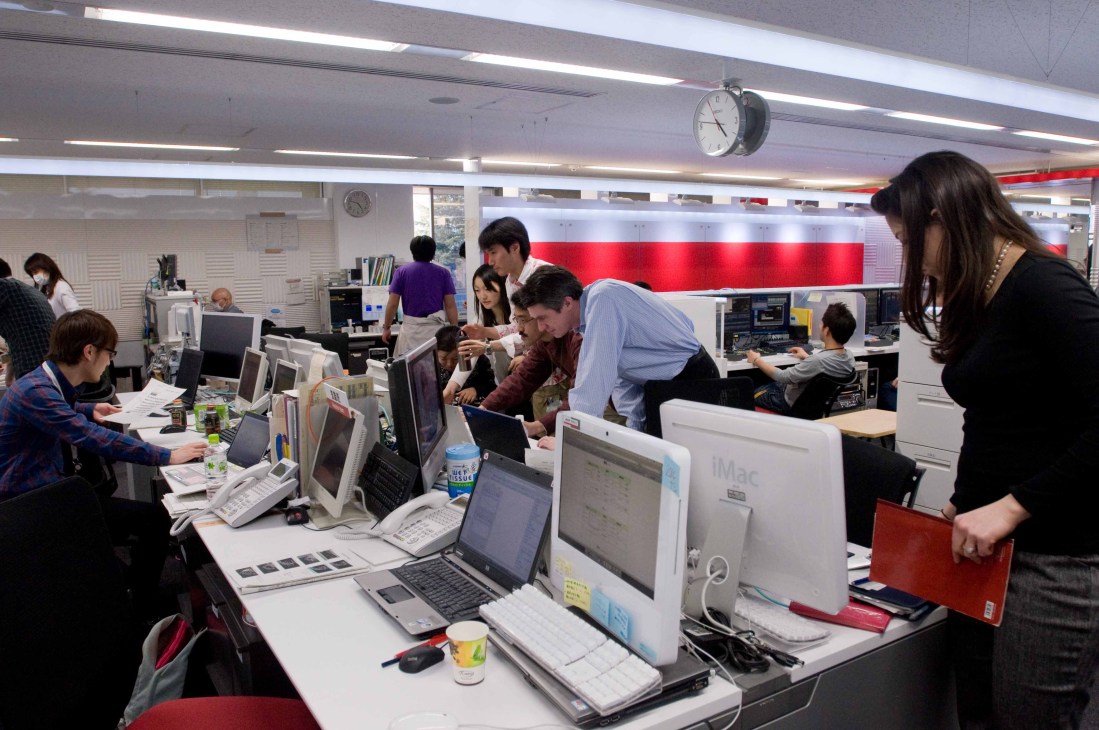 NHK World newsroom with staff working at computers and desks with red and white signage visible.