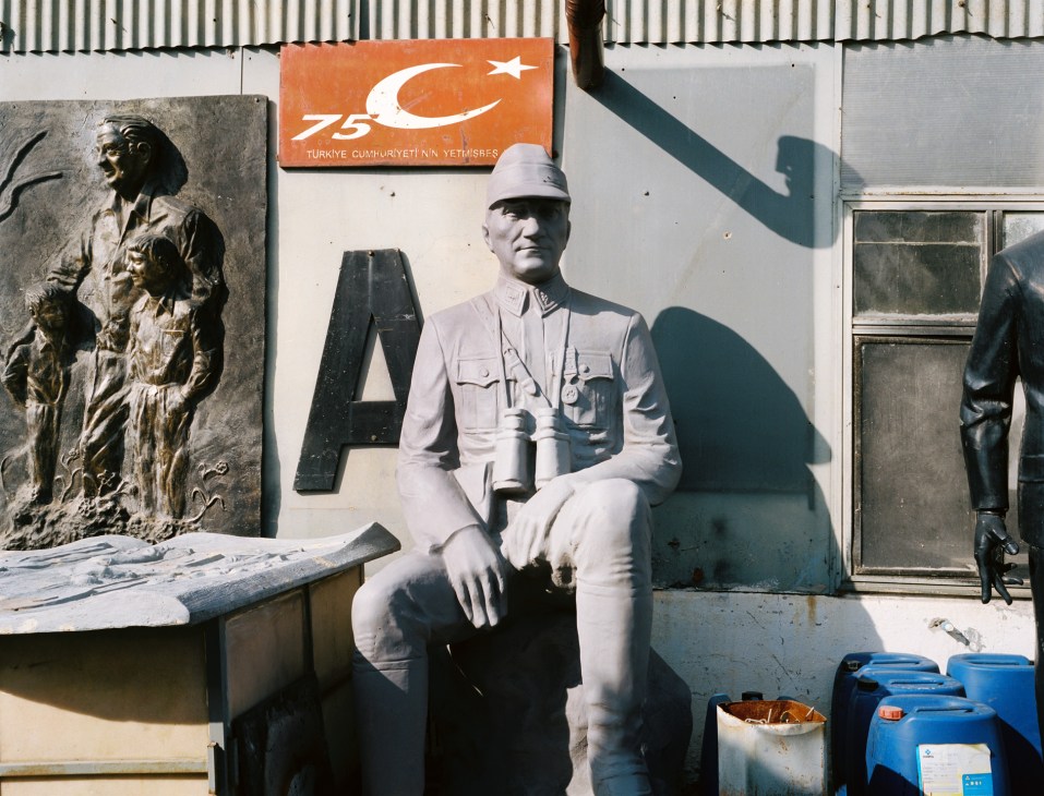 White statue of Mustafa Kemal Ataturk in military uniform holding binoculars, displayed alongside bronze relief and Turkish flag.