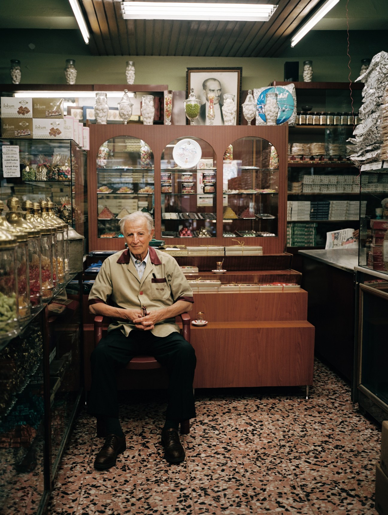 An elderly man sits in a sweet shop surrounded by glass display cases, with a framed portrait of Ataturk hanging on the wall behind him.