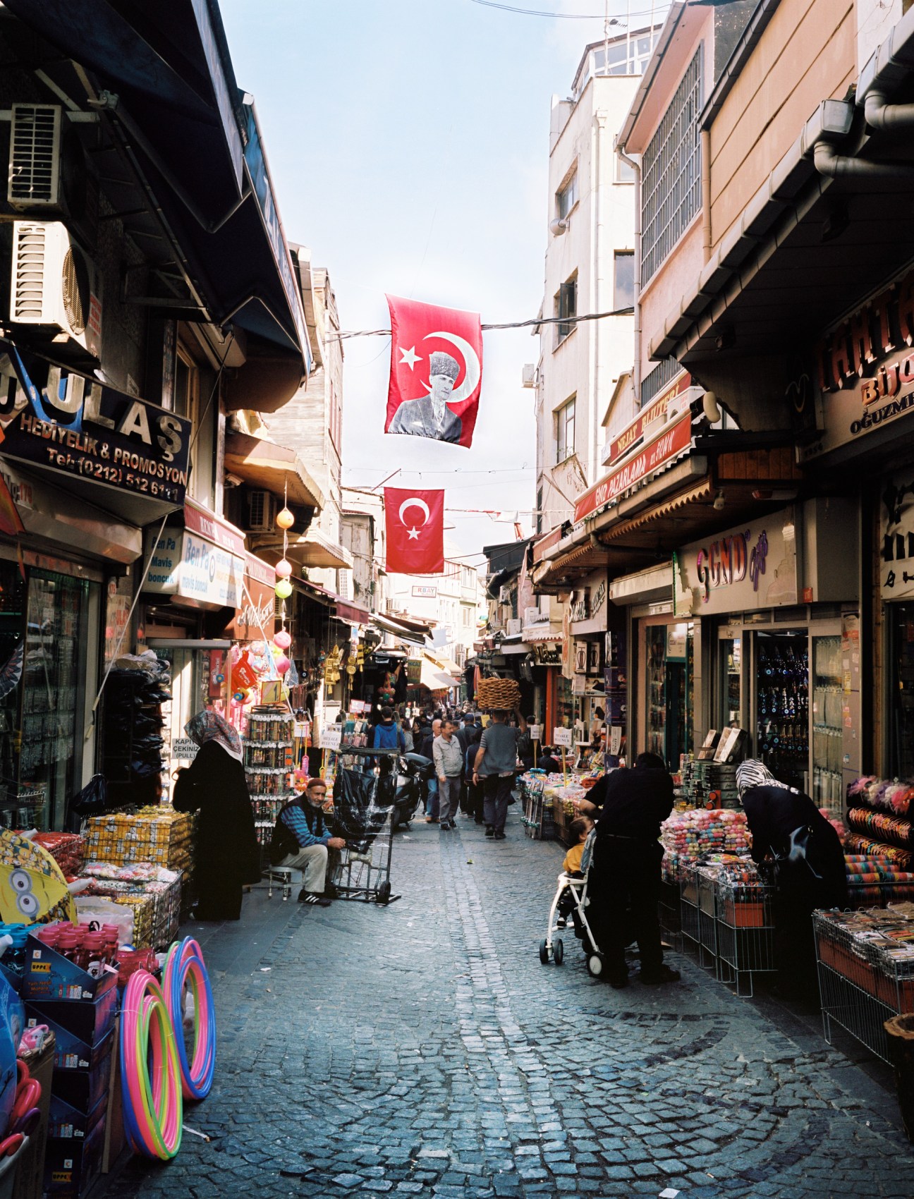 Turkish marketplace street with shops and vendors, flags featuring Ataturk's portrait hanging overhead.