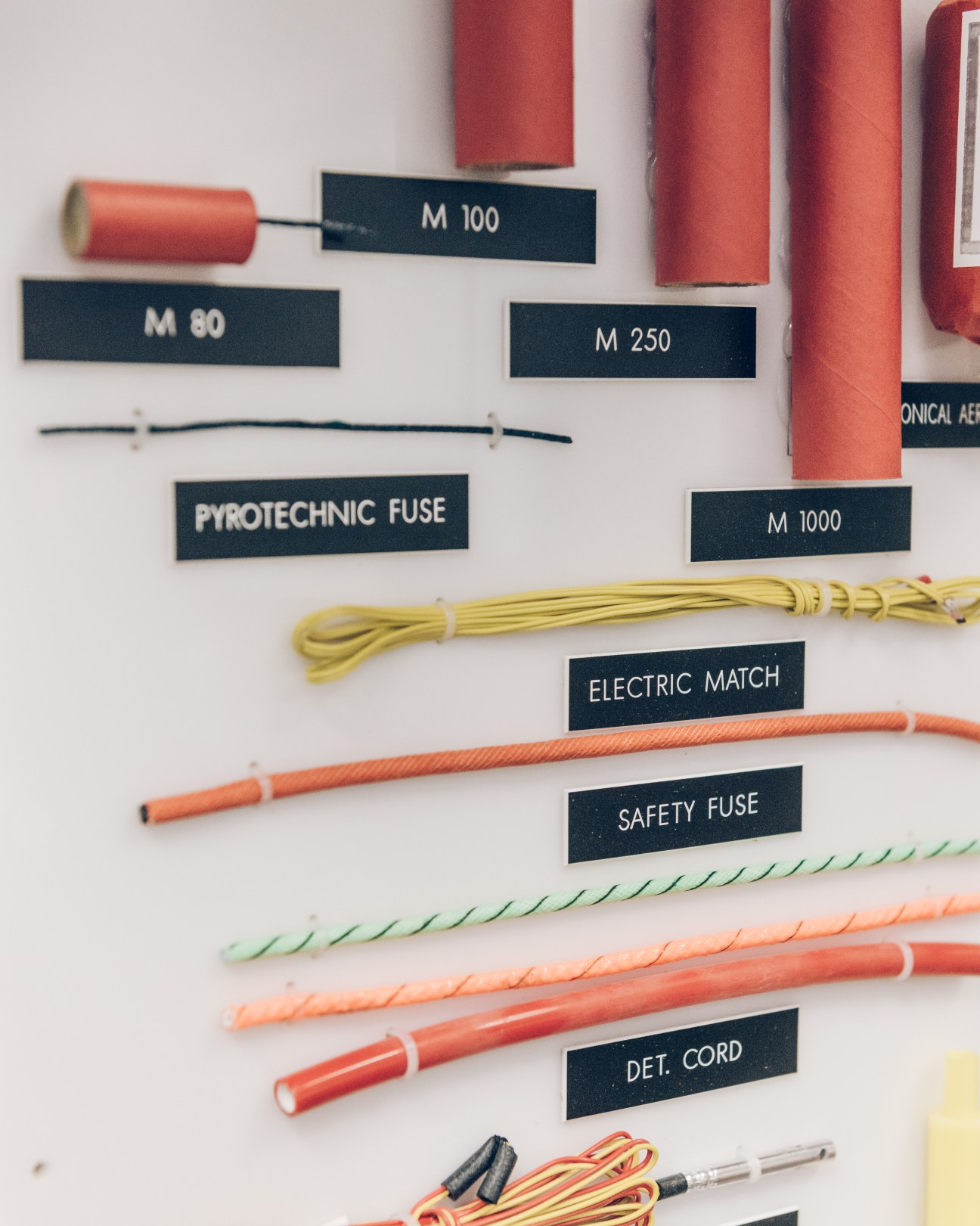 Display of various explosive devices and detonation components labeled M 80, M 100, M 250, M 1000, pyrotechnic fuse, electric match, safety fuse, and det cord.