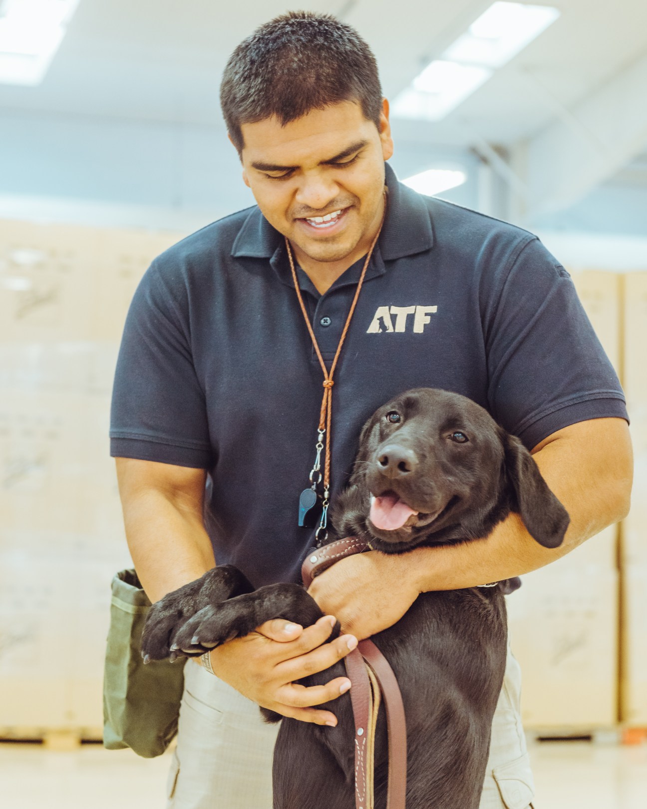 ATF handler smiling while holding a brown labrador dog indoors.