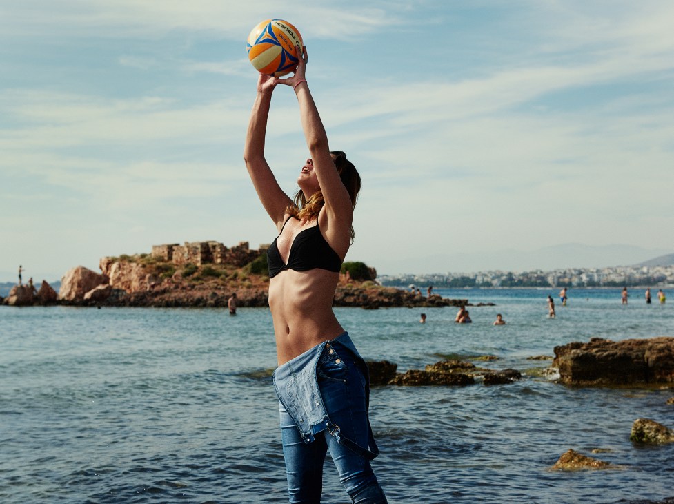 Person holding a volleyball above their head while standing in the sea at a Greek beach.