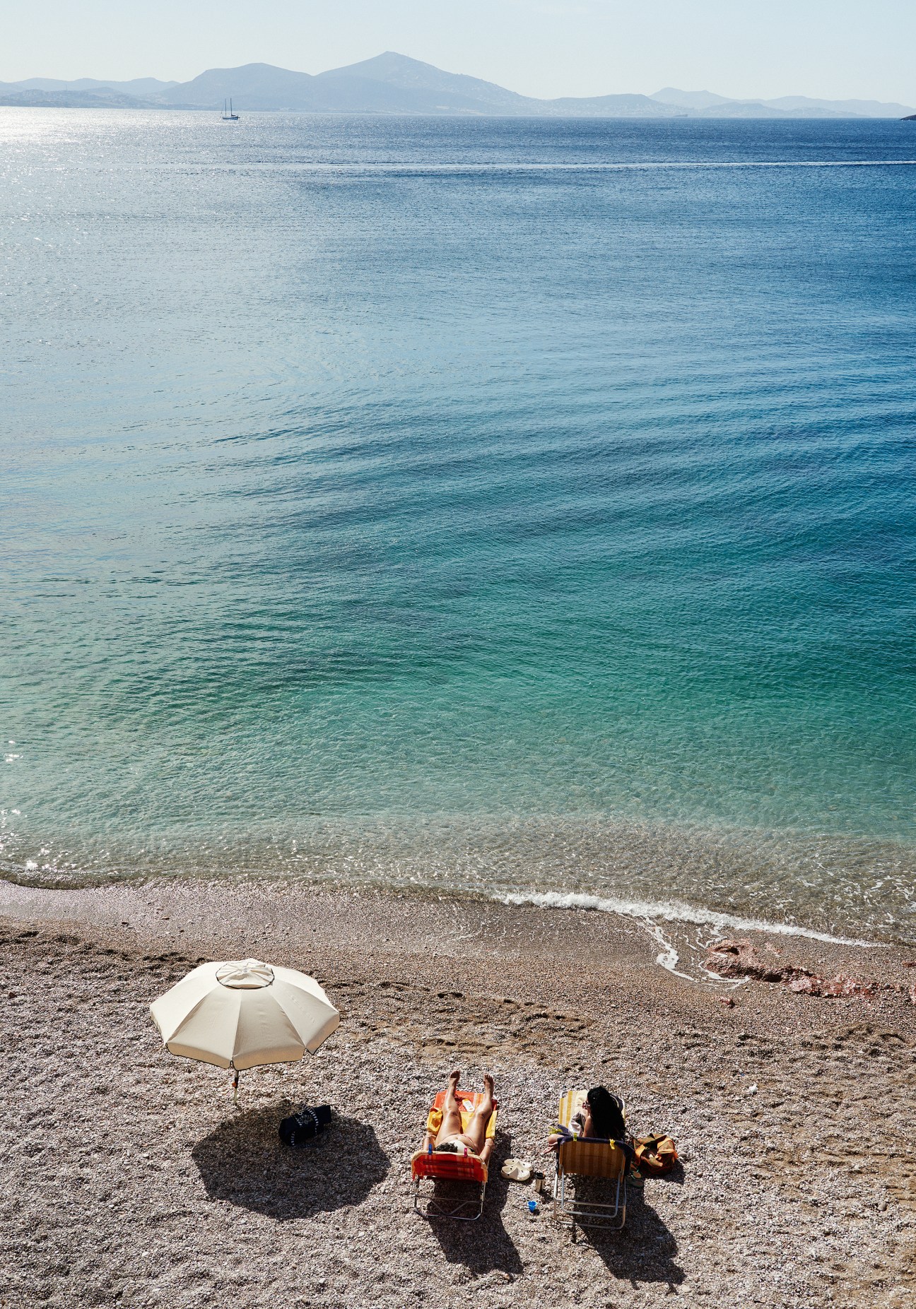 Two people relax on the sand at Varkiza beach with a white umbrella, clear turquoise waters, and mountains visible across the Aegean Sea.
