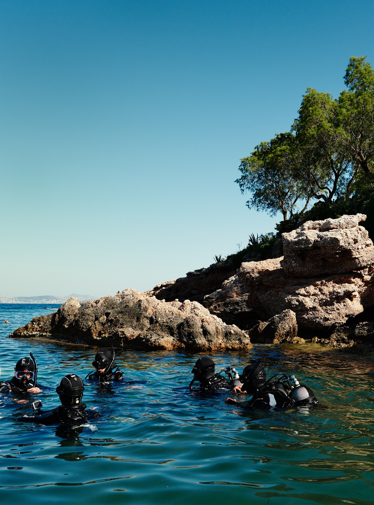 Scuba divers in wetsuits in clear blue water near rocky outcrops and vegetation in Varkiza bay, Athens.