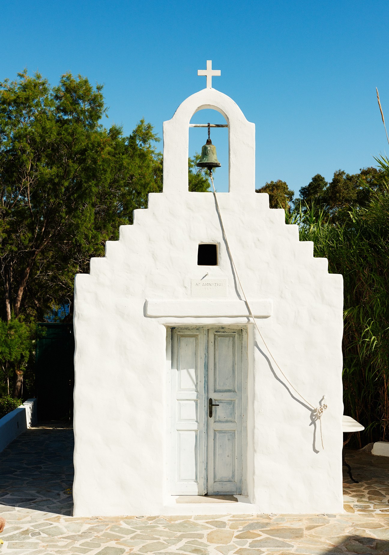 White Orthodox chapel with cross and bell in Varkiza, Greece, surrounded by green vegetation.
