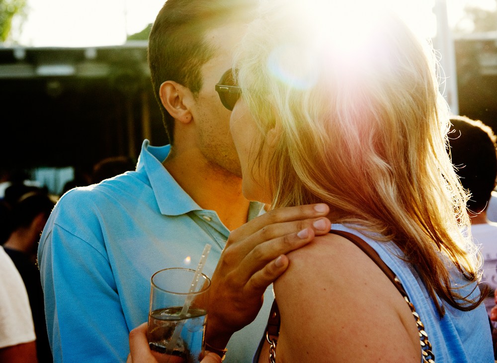 A man and woman embrace while holding drinks at a beach club gathering in golden sunlight.