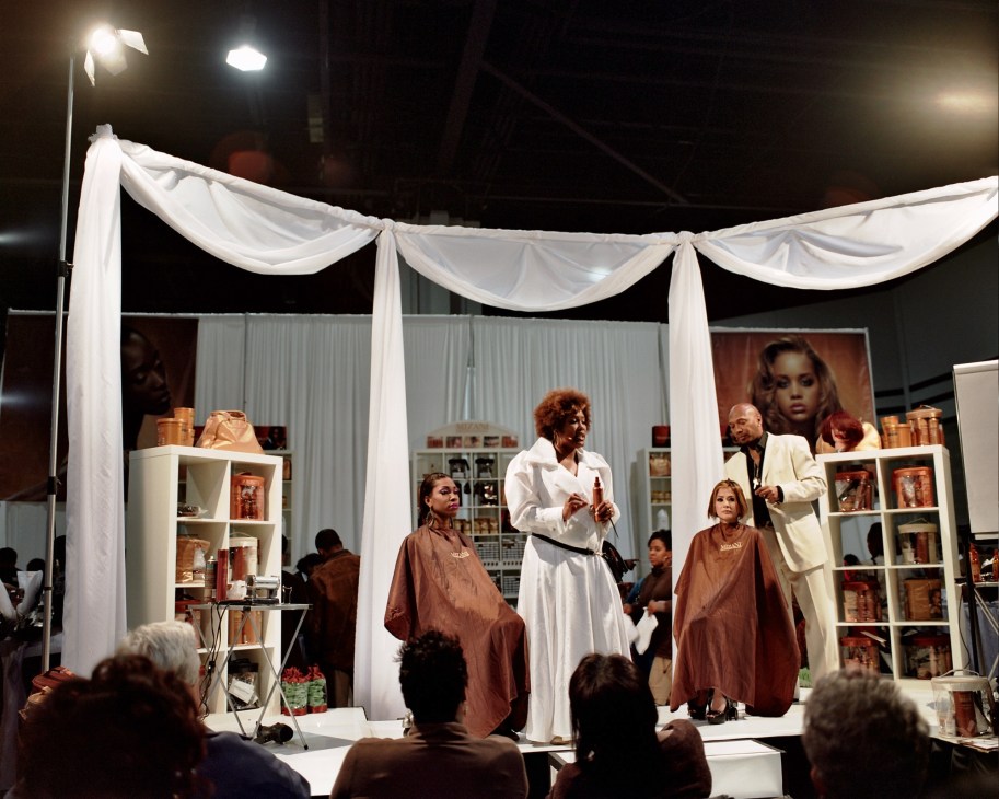 Hair stylist demonstrates weaving technique on stage under white drapes at Bronner Brothers hair show.