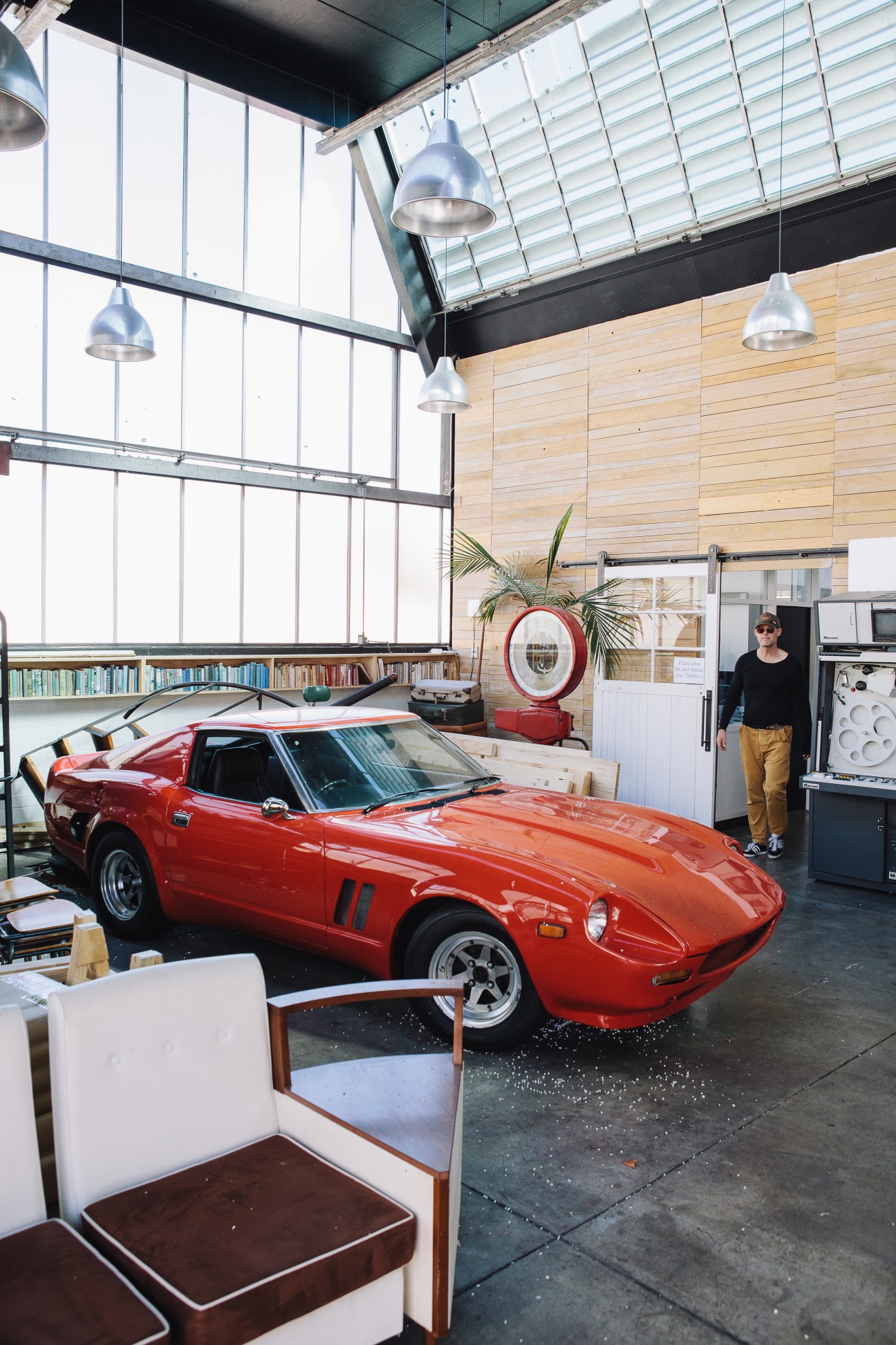 A red Datsun sports car parked inside a high-ceilinged studio space with industrial pendant lights and wooden wall paneling.
