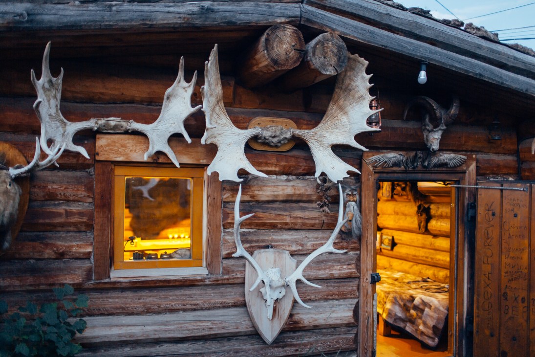 Wooden log cabin with large bleached moose antlers mounted on facade and warm golden light from windows.