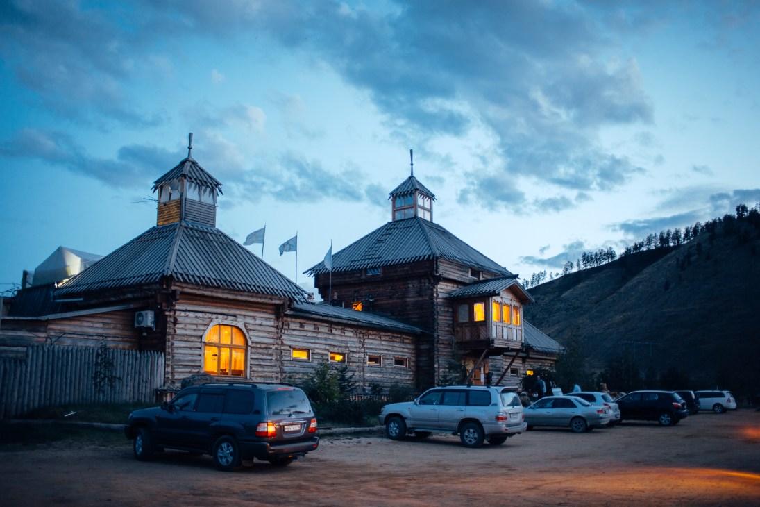 Wooden lodge with distinctive cupolas at dusk, parked cars in foreground, forested hillside behind.