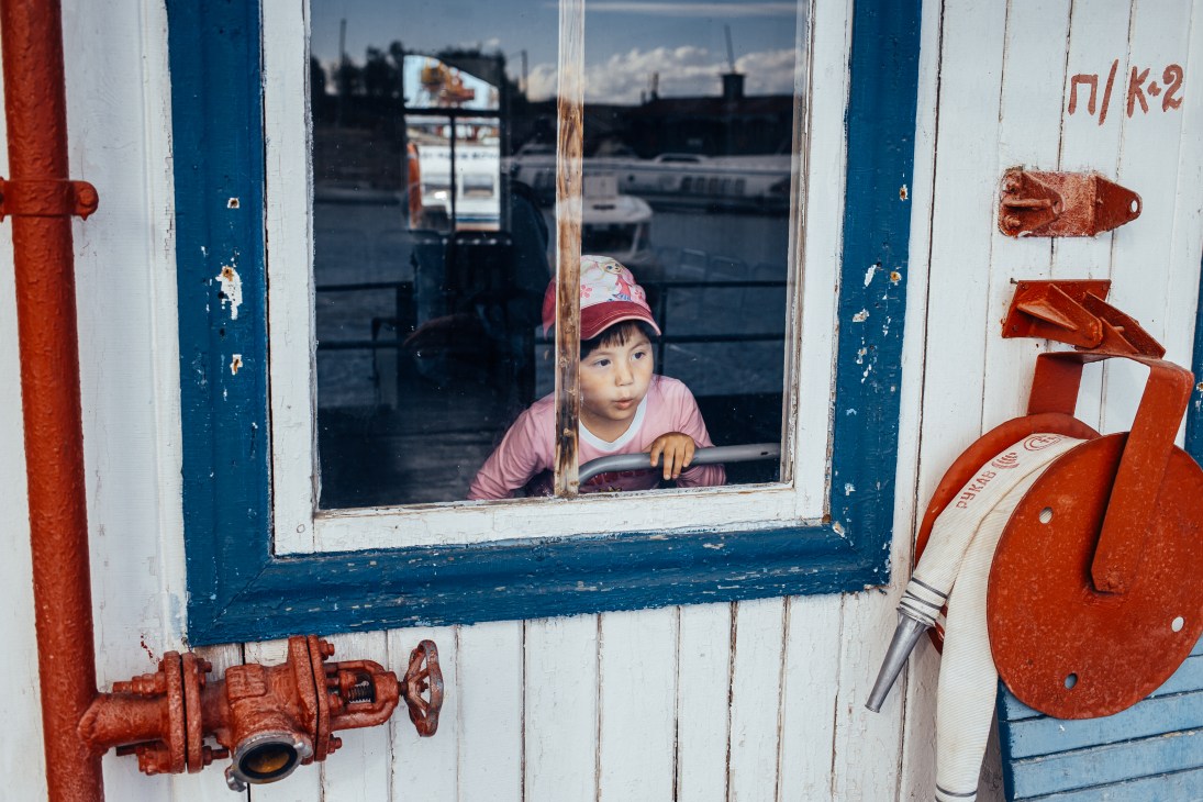 Child looking out window of a river station boat with red and blue painted exterior