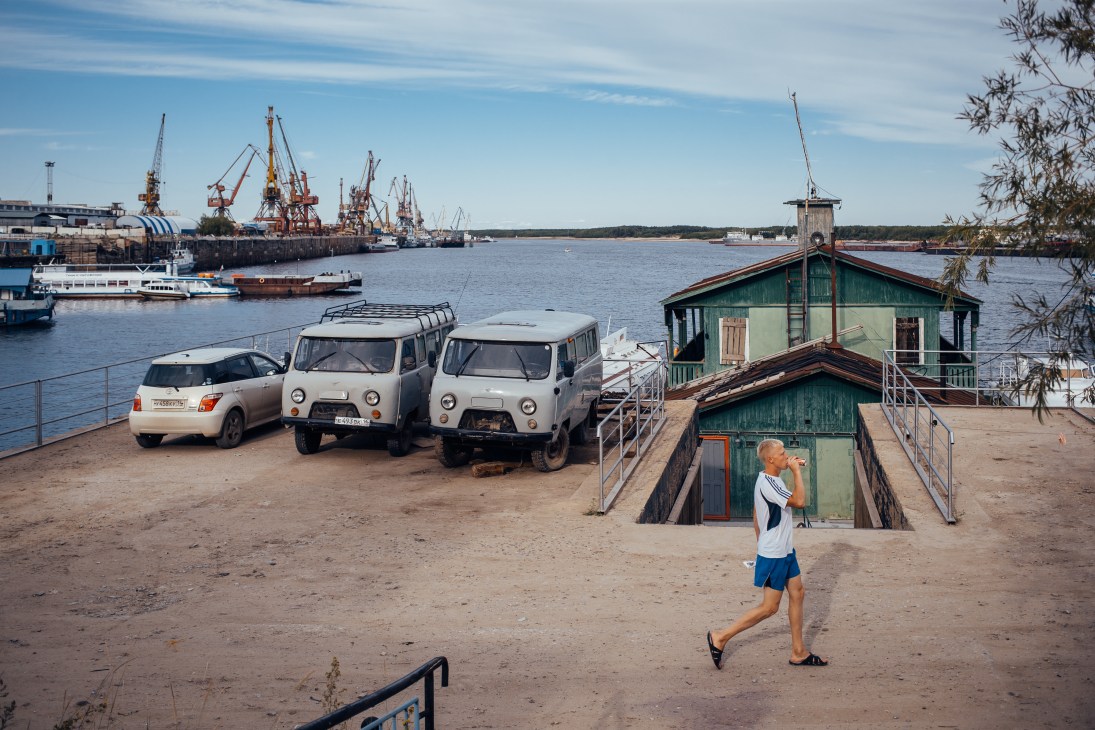 A jogger runs past parked vehicles and a green wooden structure at Yakutsk's river station with port cranes visible across the water.