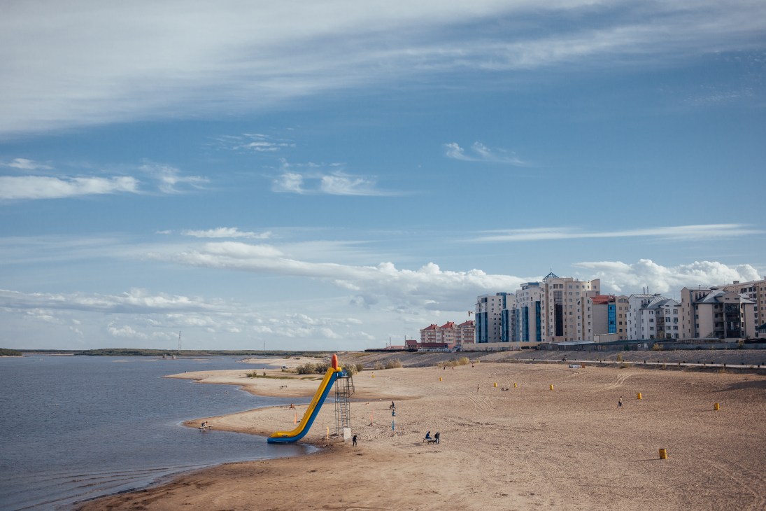 Yakutsk city beach with a large yellow-blue water slide, sandy shore, and modern apartment buildings in the background along the Lena River.