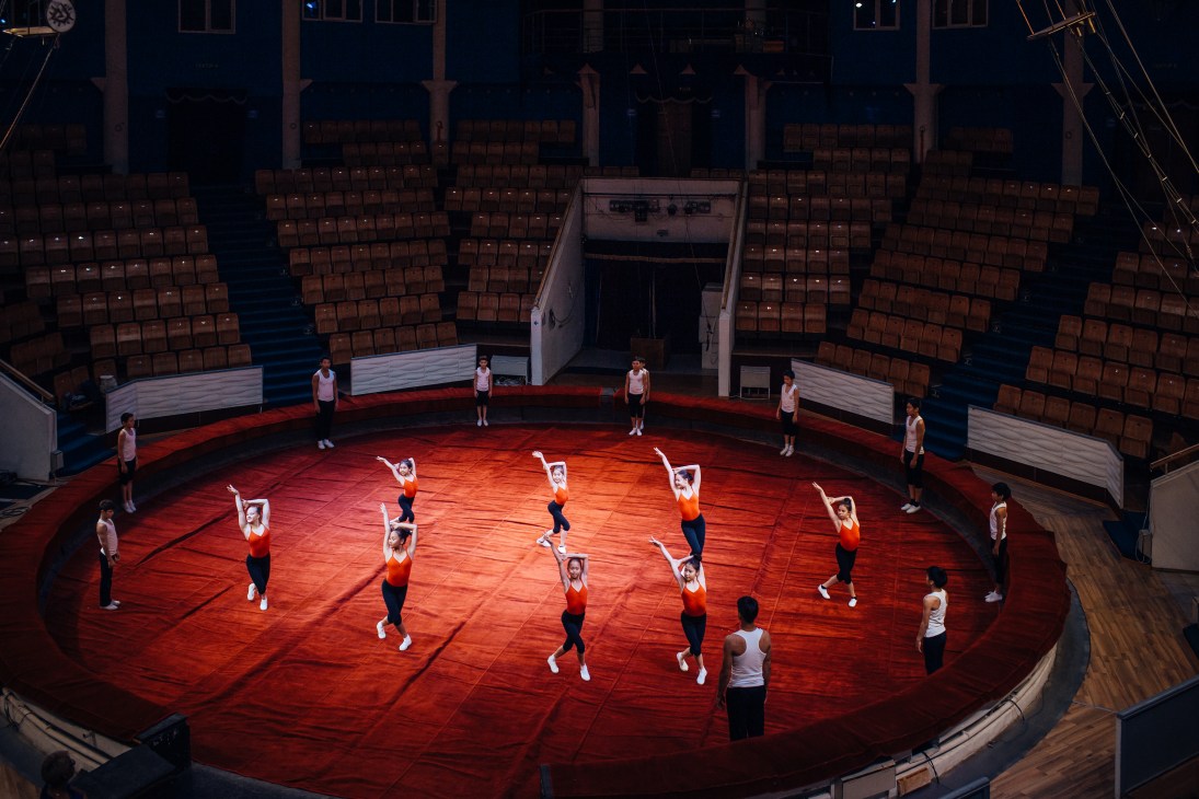 Performers in red and white athletic wear practice synchronized movements on the red circular floor of a circus ring surrounded by empty seating.