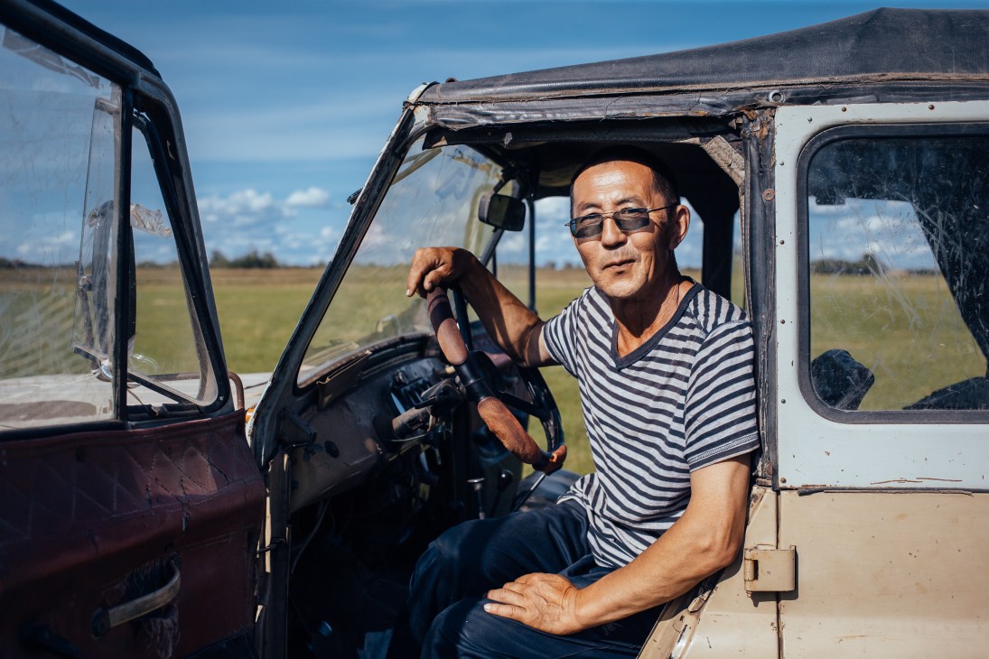 Man in striped shirt and sunglasses sits in vintage Soviet UAZ jeep in open grassland.
