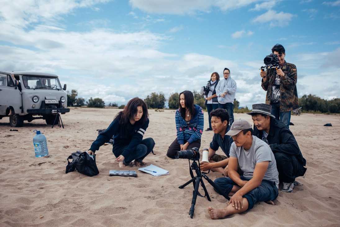 Film crew and actors on sandy riverbank with vehicle and equipment during production on Lena River