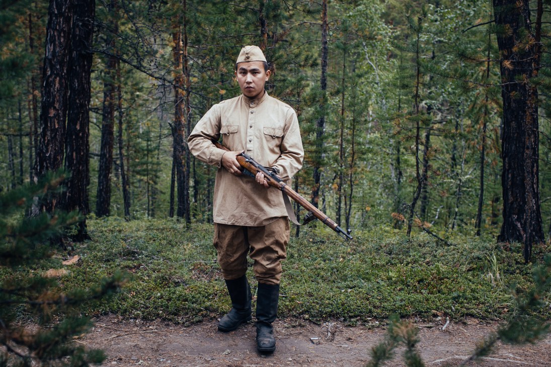 Actor in period military uniform holding rifle stands in forest during Second World War film production.